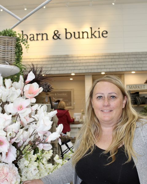 A woman stands beside flowers. On the wall behind her, are the words "barn and bunkie".