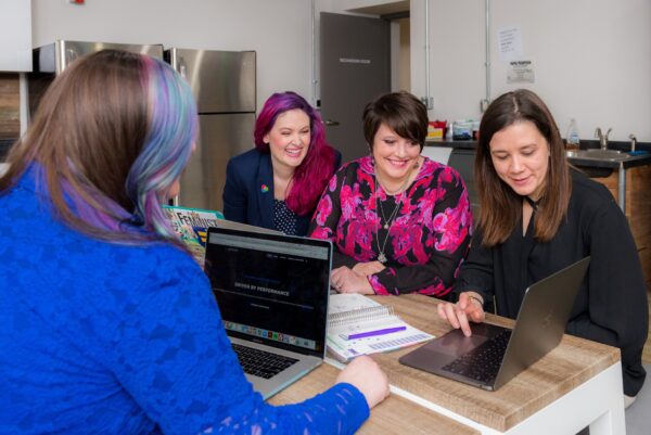 Women gather around a laptop on a table. One of these women is demonstrating something to them using the laptop.