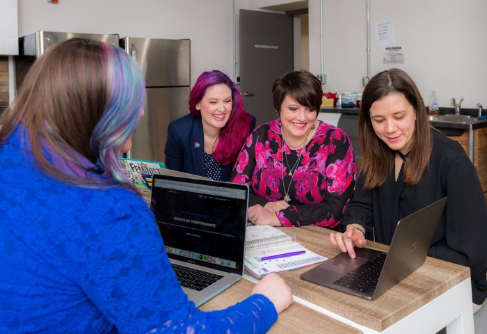 Women gather around a laptop on a table. One of these women is demonstrating something to them using the laptop.